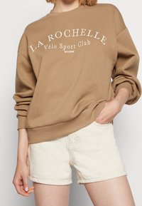 Osoba v hnědé mikině s nápisem "La Rochelle Vélo Sport Club est. 1998" a světle béžových šortkách, s rukou v kapse.