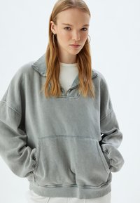 Sudadera gris claro, con tela suave, cuello con media cremallera, ajuste holgado, puños acanalados, bolsillo frontal, acabado ligeramente texturizado.