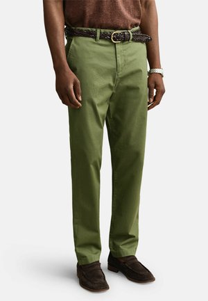 Man wearing olive green pants, brown braided belt, brown suede loafers, and a silver wristwatch against a plain background.