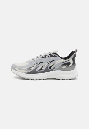 Zapatilla deportiva gris claro con patrones negros y blancos en forma de llama, suela blanca y forro interior negro, mostrada de perfil.