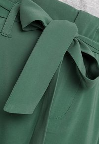Pantaloni di tessuto verde con una cintura larga annodata. Il materiale è liscio con una leggera lucentezza e c'è una tasca visibile di lato.