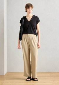 Top nero a maniche corte e scollo a V, pantaloni beige a gamba larga e sandali neri scivolati. La modella porta una borsa nera. Tessuto morbido, vestibilità ampia.