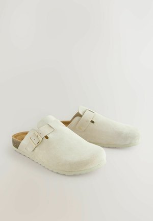 Balti zomšiniai klumpiai su lygiu paviršiumi, apvalia nosimi, reguliuojamu dirželiu ir lengvu, paminkštintu padu. Minimalistinis dizainas be raštų.