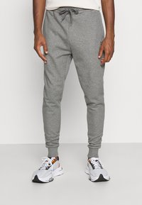 Man som bär ljusgrå joggingsbyxor med dragsko och vita, svarta och oranga sportskor, stående mot en enkel ljus bakgrund.