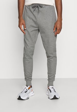 Mann in hellgrauen Jogginghosen mit Kordelzug und weißen, schwarzen und orangefarbenen Sportschuhen, der vor einem schlichten hellen Hintergrund steht.