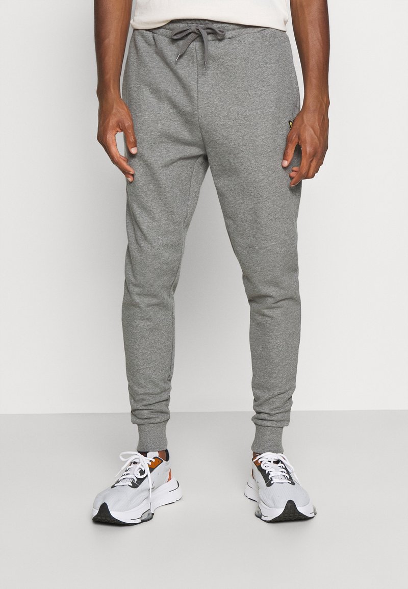 Man som bär ljusgrå joggingsbyxor med dragsko och vita, svarta och oranga sportskor, stående mot en enkel ljus bakgrund.