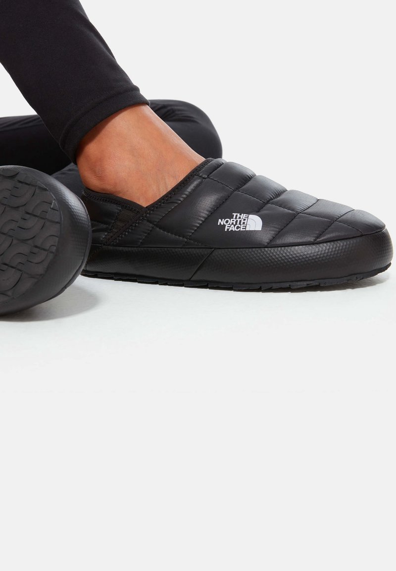 Personne portant des chaussons noirs matelassés à enfiler avec le logo The North Face, assise avec une jambe pliée et l'autre étendue.