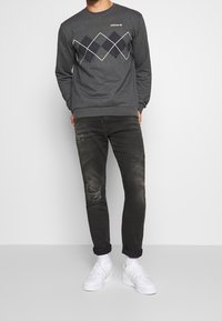 Grå sweatshirt med argyle-mönster i mörkblått, kombinerad med svarta slitna jeans och vita sneakers, vilket skapar en avslappnad outfit.