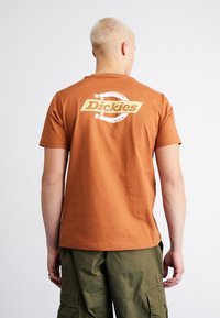 Camiseta de algodón marrón con mangas cortas, que presenta un logo "Dickies" en blanco y amarillo en la parte posterior, combinada con pantalones cargo verde oliva.