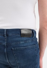 Ciemnoniebieskie dżinsy o slim fit, z skórzaną naszywką marki z tyłu w pasie i szczegółowym przeszyciem na kieszeniach.