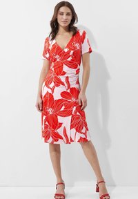 Rotes Blumenkleid bis zu den Knien mit kurzen Ärmeln und V-Ausschnitt, auf einem pinken Hintergrund und mit tailliertem Schnitt, ergänzt durch rote Sandalen.
