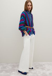Cárdigan de punto de gran tamaño de múltiples colores con rayas azules, verdes y rosas, llevado sobre una camisa de mezclilla, combinado con pantalones anchos blancos y zapatos azules.