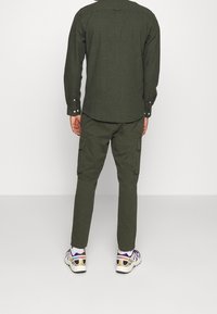 Man i olivgrön långärmad skjorta och cargobyxor med vita sneakers, står mot en enkel vit bakgrund, ryggen synlig.