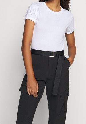 Femme portant un t-shirt blanc uni à manches courtes rentré dans un pantalon noir taille haute avec une ceinture noire et des poches latérales.