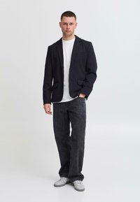 Veste blazer bleu marine foncé sur un T-shirt blanc, associée à un jean gris foncé à jambes larges. Le modèle porte des baskets grises au design minimaliste et au tissu lisse.