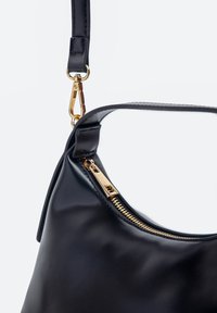 Schwarze Lederhandtasche mit goldenem Reißverschluss und abnehmbarem Riemenverschluss vor schlichtem hellem Hintergrund.