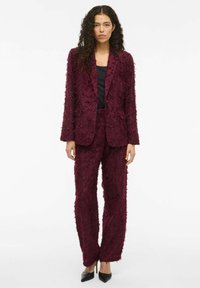 Burgundský strukturální oblek s květinovou látkou, obsahující přiléhavý blazer a rovné kalhoty. Černý top a špičaté podpatky dotvářejí vzhled.