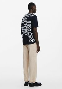 Camiseta gráfica marina con patrones de texto en blanco bold; combinada con pantalones cargo beige y zapatos negros. Fondo liso.