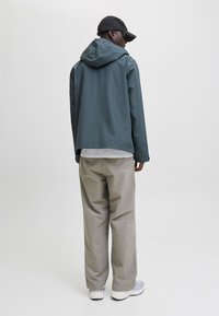 Mand iført en mørkegrå hættetrøje, beige vidde benbukser, hvide sneakers og en sort baseballkasket, stående med fronten væk fra kameraet mod en ensfarvet baggrund.