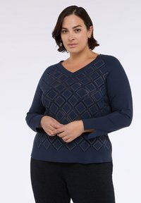 Námořnický modrý svetr s výstřihem do V a dlouhými rukávy. Obsahuje diamantový vzor s výřezy a malými ozdobami po celé přední části.