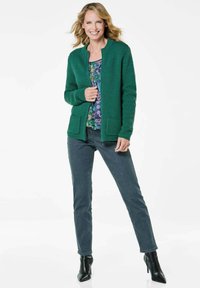 Grüne Strickjacke mit strukturiertem Muster, zwei Fronttaschen, über einem blumigen Oberteil getragen. Kombiniert mit dunklen Jeans und spitzen schwarzen Stiefeln.
