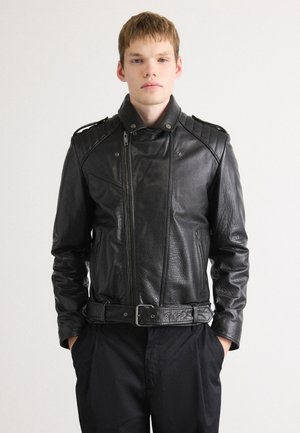 Young man wearing a black leather biker jacket with padded shoulders and a belt, standing with hands in black trousers pockets.