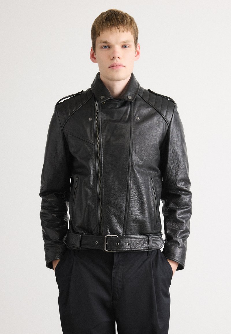 Young man wearing a black leather biker jacket with padded shoulders and a belt, standing with hands in black trousers pockets.