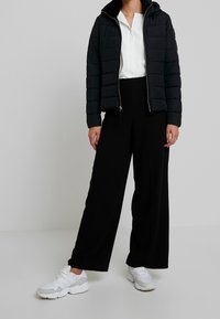 Polstret sort jakke med lynlås, hvid bluse og sorte vide bukser. Hvide sneakers med grå detaljer. Minimalistisk design.