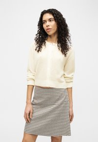 Γυναίκα με μακριά σγουρά μαλλιά που φοράει εκρού ζακέτα με κουμπιά και μπεζ-μαύρη καρό φούστα, που στέκεται μπροστά σε απλό φόντο.