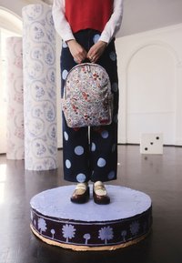 Személy áll festett platformon, kezében mintás hátizsák, piros pulóvert, pöttyös nadrágot és krémszínű, barna cipőt visel világos szobában.