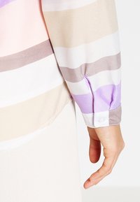 Bras droit et main portant une chemise à manches longues avec des rayures horizontales beige, blanches, marron et violettes et un poignet boutonné.