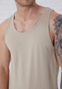 Beige tank top lavet af blød stof, med rund halsudskæring og ærmeløs design. Synlige tatoveringer på armen tilføjer detaljer.