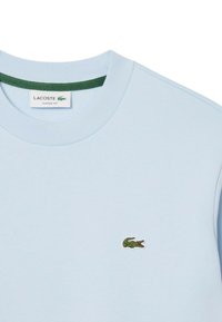 Sudadera de algodón azul claro con cuello redondo, con un acento verde en el cuello y un pequeño logotipo de cocodrilo bordado en verde.