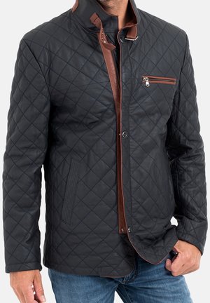 Mann trägt schwarze gesteppte Jacke mit braunen Lederbesätzen, Reißverschlusstasche an der Brust und Fronttaschen, kombiniert mit blauen Jeans.