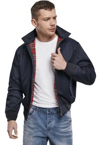 Veste de bombardier navy avec une doublure écossaise rouge, fermeture éclair, poignets côtelés et deux poches latérales. Portée par-dessus un t-shirt blanc.