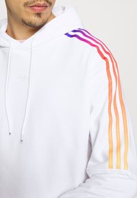 Man met een witte hoodie met paarse, roze, oranje en gele gradient strepen op de schouder en bovenarm, gedeeltelijk zichtbare gezicht.