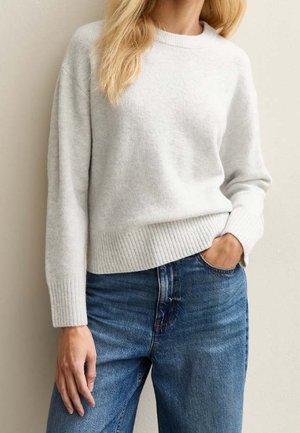 Personne portant un pull gris clair à manches longues et un jean bleu avec une main dans la poche, sur un fond uni.