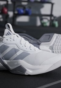 Scarpe sportive Adidas bianche e grigie con suole testurizzate posizionate su un pavimento scuro di una palestra con kettlebell sfocati sullo sfondo.