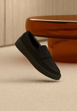 Sort slip-on loafer med tykk såle hengt opp over beige teppe, med et moderne brunt rundt bord i bakgrunnen.