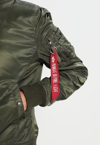 Zielona kurtka bombowa wykonana z błyszczącego nylonu, z prążkowanymi mankietami i kieszenią na suwak na rękawie, z czerwoną zawieszką z napisem "USUNĄĆ PRZED LOTEM."