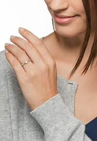 Anello d'argento con una pietra preziosa rotonda centrale, banda sottile, indossato su una mano. Sullo sfondo si vede un maglione lavorato a maglia grigio chiaro.