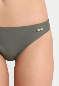 Olivegrüner Bikiniunterteil mit glatter Textur, minimalistischem Nahtdesign und einem kleinen silbernen Logoakzent an der Seite.
