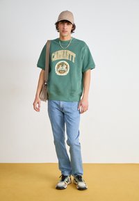 Zelená mikina s zlatým logem "Carhartt", světlé modré džíny, béžová kšiltovka a multibarevné tenisky. Neutrální pozadí se žlutou podlahou.