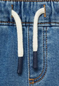 Denimtyg med en blå ton, med en dragsko med flätad vit struktur och marinblå ändar. Synliga sömnadsdetaljer ger extra karaktär.