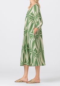 Green patterned dress with a loose fit, knee-length cut, three-quarter sleeves, and pockets. Worn with strappy beige sandals.