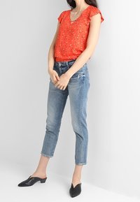 Rode blouse met korte mouwen en een V-hals, versierd met kleine geometrische patronen, gecombineerd met lichtblauwe cropped jeans en zwarte puntschoenen.