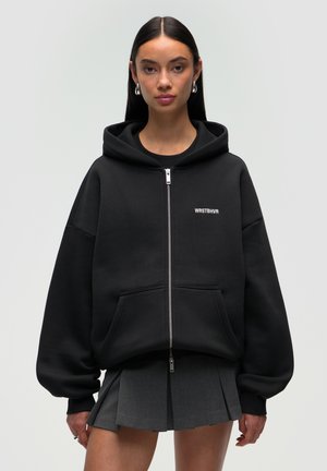 Schwarzer Reißverschluss-Hoodie mit großzügigem, lockeren Schnitt, Fronttaschen und gesticktem "WRSTBHVR"-Logo. Begleitet von einem grauen plissierten Rock.