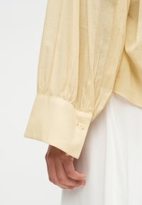 Manche beige à manches longues avec poignet froncé et fermeture à boutons sur le bras d'un mannequin, portant un vêtement blanc sous la manche.