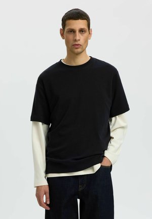 Uomo con capelli corti e scuri che indossa una camicia nera a maniche corte sopra una camicia bianca a maniche lunghe e jeans scuri, in piedi davanti a uno sfondo chiaro e uniforme.
