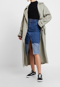 Femme portant un long trench-coat beige, un haut noir côtelé, une jupe crayon en denim bicolore avec une fente à l'avant, et des baskets noir et blanc.